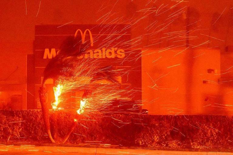 A imagem mostra um incêndio em um arbusto， com chamas e faíscas voando em um ambiente de luz avermelhada. Ao fundo， é visível o letreiro de um restaurante McDonald039;s， que está iluminado pela luz do fogo. O céu parece estar coberto de fumaça， contribuindo para a atmosfera dramática da cena.