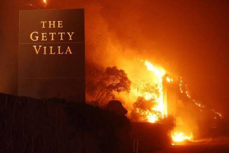 A imagem mostra um sinal iluminado que diz 039;THE GETTY VILLA039; em meio a um incêndio florestal. Chamas e fumaça são visíveis ao fundo， criando uma atmosfera de emergência. A cena é noturna， com um brilho laranja intenso das chamas contrastando com a escuridão ao redor.