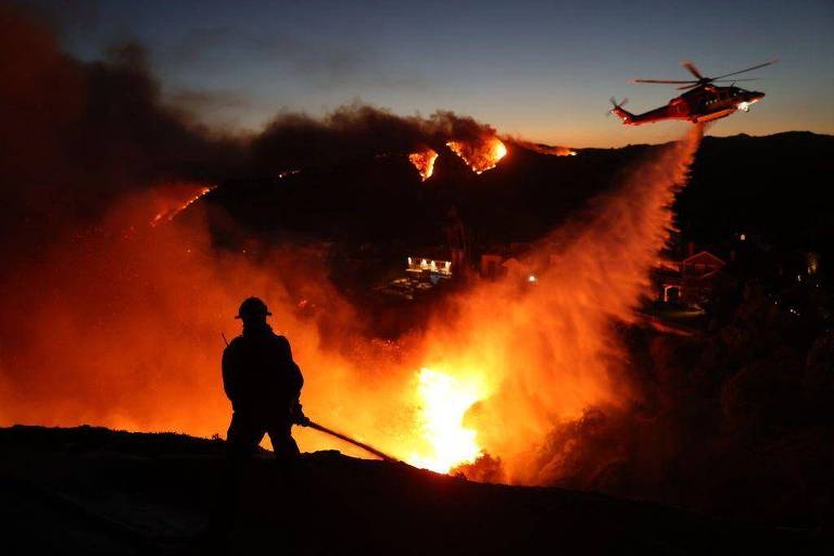 A imagem mostra um bombeiro em silhueta， observando um incêndio florestal. No fundo， chamas intensas iluminam a cena， enquanto um helicóptero lança água sobre o fogo. O céu está escuro， com um gradiente de cores que vai do azul ao laranja devido ao incêndio.