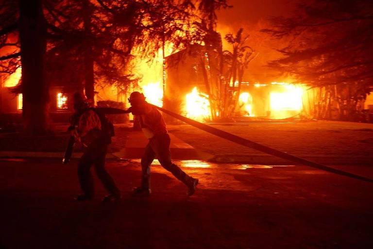 A imagem mostra dois bombeiros correndo em direção a um incêndio. O fundo é dominado por chamas intensas e fumaça， iluminando a cena com uma luz laranja e vermelha. Os bombeiros estão usando equipamentos de proteção e carregando uma mangueira de incêndio.