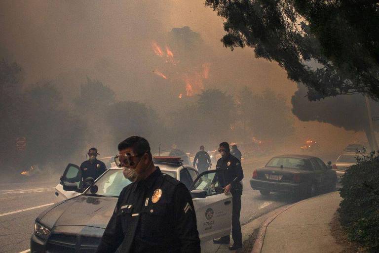 A imagem mostra policiais em uma cena de incêndio florestal. Eles estão usando máscaras e estão próximos a viaturas policiais. Ao fundo， há fumaça densa e chamas visíveis entre as árvores. A estrada está parcialmente visível， com outros veículos estacionados.