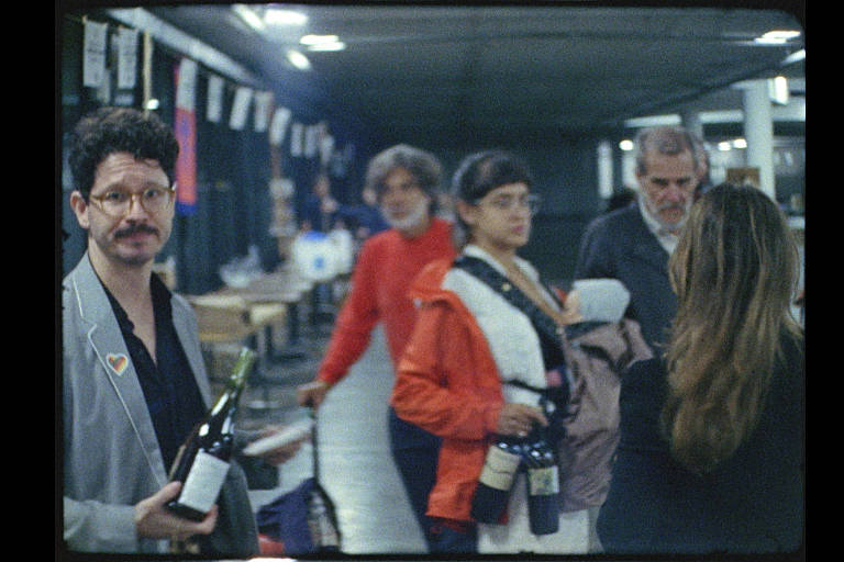 A imagem mostra um grupo de pessoas em um ambiente interno， possivelmente em um evento social. Um homem à esquerda segura uma garrafa de vinho e sorri para a câmera. Ao fundo， há várias pessoas conversando， algumas usando roupas coloridas， como uma mulher vestindo uma jaqueta vermelha. O ambiente parece ser bem iluminado， com mesas e cadeiras visíveis.