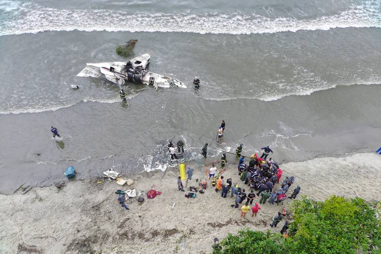 A imagem mostra uma cena de um acidente na praia, onde um barco está encalhado na água. Há várias pessoas na areia e na água, algumas parecendo ajudar. O ambiente é de praia, com ondas e vegetação ao redor.