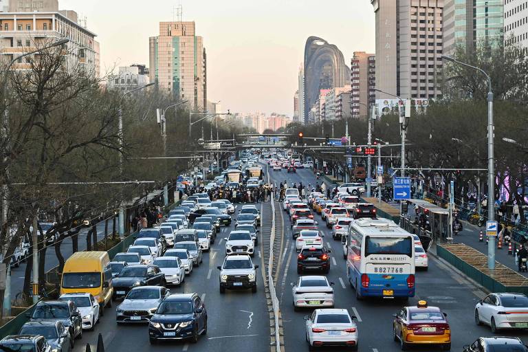 A imagem mostra uma rua movimentada em uma cidade， com muitos carros em diferentes direções. Há prédios altos ao fundo， e a iluminação sugere que é no final da tarde. Árvores estão visíveis ao longo da calçada， e há sinais de trânsito e postes de luz ao longo da via.