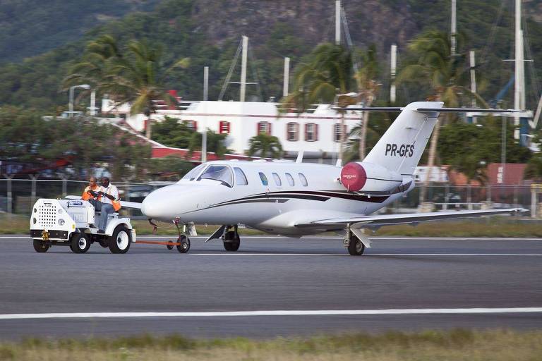 Um avião a jato pequeno está sendo rebocado por um veículo de serviço na pista de um aeroporto. Ao fundo， há edifícios e árvores， sugerindo um ambiente tropical.