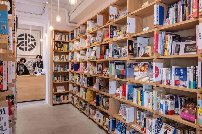 A imagem mostra o interior de uma livraria com prateleiras de madeira， em três lados， repletas de livros organizados， com alguns títulos visíveis， muitos deles em caracteres japoneses. No fundo， duas pessoas estão em um balcão de atendimento， ambas com cabelo liso escuro， usando quimonos pretos em que se pode ler， em branco， o nome da livraria， Honmaru. O ambiente é iluminado por lâmpadas pendentes. O piso é claro.