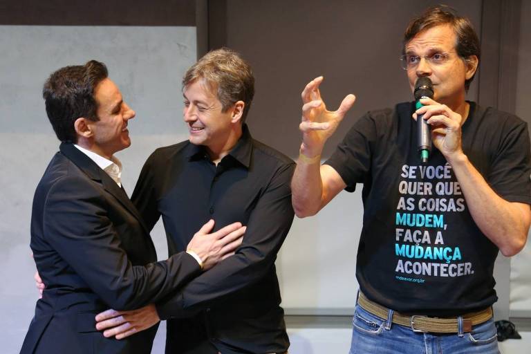 A imagem mostra três homens em um evento. O homem à esquerda， vestido de terno escuro， está abraçando o homem do meio， que usa uma camisa clara. O homem à direita， que está falando ao microfone， veste uma camiseta preta com texto em letras brancas. O ambiente parece ser uma sala de conferências ou evento.