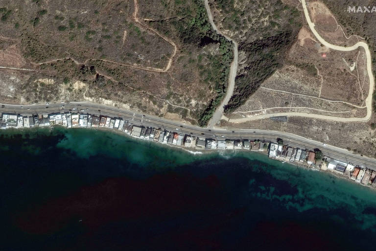 A imagem mostra uma vista aérea de uma costa, onde uma estrada se estende ao longo da linha da água. Há várias construções próximas à praia, com um fundo de vegetação e terreno irregular. A água é de um tom azul profundo, e a área ao redor parece ser predominantemente árida.