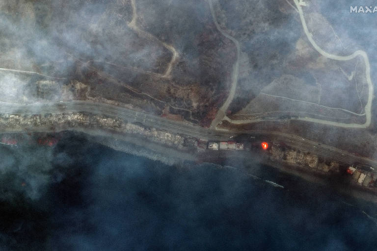 A imagem mostra uma vista aérea de uma área afetada por incêndio florestal. É possível ver uma estrada que contorna a área, com fumaça densa cobrindo a região. Há focos de fogo visíveis ao longo da estrada, e a vegetação ao redor parece queimada. A água de um corpo d'água está presente na parte inferior da imagem, contrastando com a fumaça e os danos visíveis.