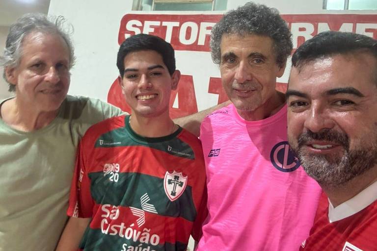 Quatro homens posando juntos em um ambiente interno. Eles estão sorrindo e vestindo camisetas de times de futebol. O homem à esquerda usa uma camiseta clara， o segundo usa uma camiseta com listras verdes e vermelhas， o terceiro está vestido com uma camiseta rosa e o último usa uma camiseta vermelha