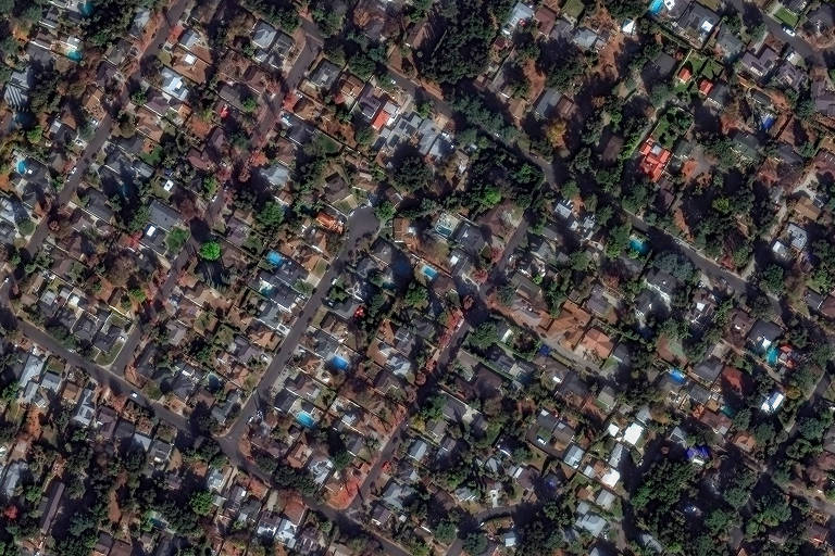 Imagem aérea de uma área residencial, mostrando uma densa disposição de casas e ruas. As casas variam em tamanho e estilo, com telhados de diferentes cores. Há áreas verdes visíveis entre as residências, sugerindo a presença de árvores e jardins. A imagem é capturada em um ângulo que permite observar a organização das ruas e a disposição das casas.