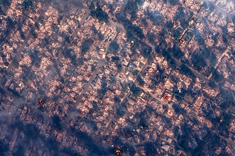 Imagem aérea mostrando uma área urbana devastada por incêndios. As casas estão visivelmente danificadas, com muitas estruturas em ruínas. Há fumaça visível cobrindo partes da imagem, indicando a presença de incêndios ativos. A vegetação ao redor também parece afetada, com áreas queimadas e desmatadas.