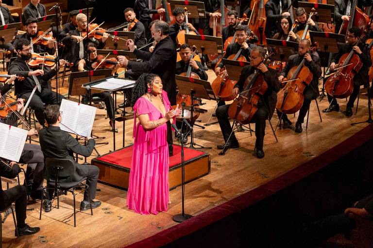 A imagem mostra uma orquestra em uma apresentação ao vivo. No centro， uma cantora vestindo um vestido rosa se apresenta em frente a um microfone. Atrás dela， uma orquestra composta por músicos tocando violinos， violoncelos e outros instrumentos. O palco é iluminado e há partituras visíveis em suportes. O ambiente é de um auditório com uma plateia visível à direita.