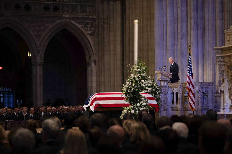 A imagem mostra uma cerimônia fúnebre em um local religioso. Um caixão coberto com a bandeira dos Estados Unidos está em primeiro plano, cercado por flores brancas. Ao fundo, um homem está em um púlpito, fazendo um discurso, enquanto uma multidão de pessoas vestidas de preto o escuta. Uma vela acesa está posicionada ao lado do caixão, e a bandeira americana está visível ao lado do orador