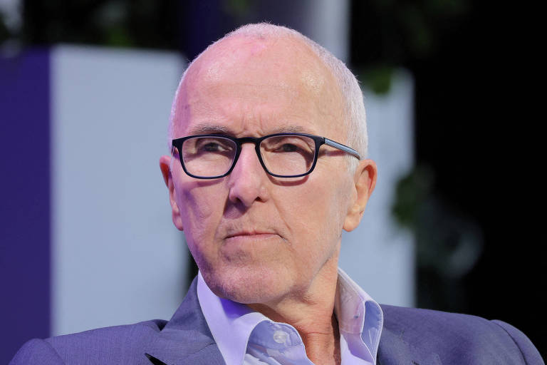 Imagem mostra Frank McCourt， um homem de cabelo calvo e pele clara， usando óculos de armação preta. Ele está com uma expressão séria e veste um paletó cinza. O fundo é desfocado， com elementos de plantas e uma iluminação suave.