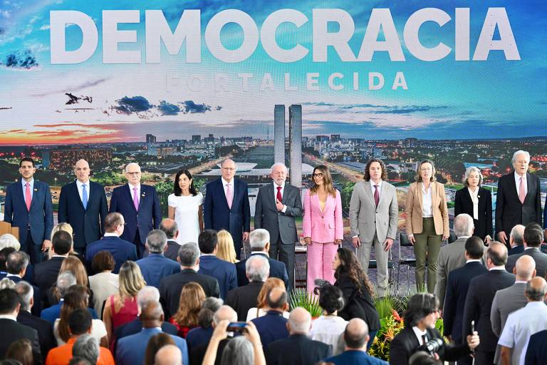 A imagem mostra um grupo de pessoas em um evento， posando em frente a um grande painel que diz 039;DEMOCRACIA FORTALECIDA039;. Ao fundo， há uma vista da cidade com prédios e um céu azul. As pessoas estão vestidas formalmente e parecem estar em um ambiente de celebração ou cerimônia.
