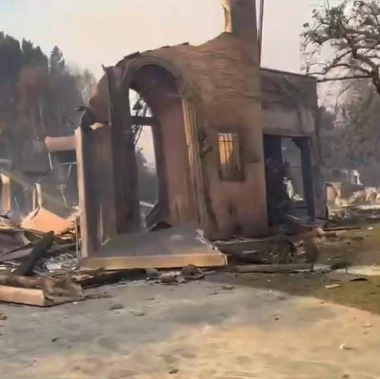 A imagem mostra uma casa parcialmente destruída por um incêndio， com paredes queimadas e uma chaminé visível. O entorno é de escombros e vegetação queimada， com árvores ao fundo. O céu parece nublado， indicando fumaça ou poluição