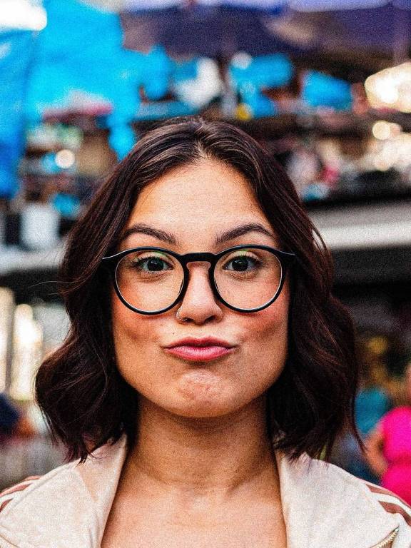 A imagem mostra uma mulher com cabelo castanho escuro e ondulado, usando óculos grandes e redondos. Ela faz uma expressão divertida, com os lábios franzidos. Ao fundo, há uma feira ou mercado com barracas e pessoas, além de um céu parcialmente coberto por lonas azuis. A iluminação é natural e a cena transmite um ambiente descontraído.