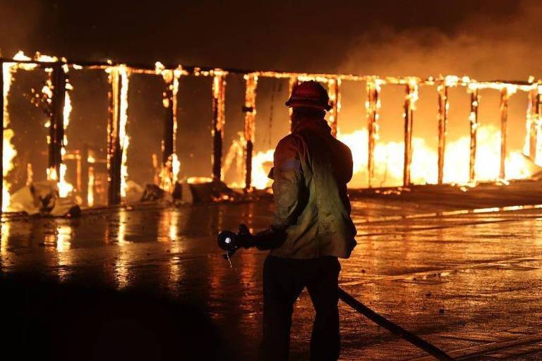 A imagem mostra um bombeiro de costas， usando um capacete e uma roupa de proteção， enquanto combate um incêndio. As chamas estão consumindo uma estrutura ao fundo， iluminando a cena com uma luz laranja intensa. O chão está molhado， refletindo a luz do fogo.