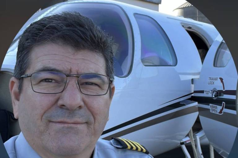 Um homem com cabelo escuro e grisalho， usando óculos e uma camisa de piloto cinza com insígnias amarelas nos ombros， está posando para a foto ao lado de uma aeronave. A parte da aeronave visível é branca com detalhes em preto e há uma porta aberta. O fundo é desfocado， mas parece ser um ambiente de aeroporto.