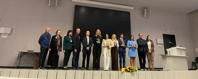 Um grupo de pessoas está posando em um palco. Ao fundo， há uma tela grande e um espaço vazio à esquerda. As pessoas estão vestidas de maneira variada， algumas em trajes formais e outras em roupas casuais. O ambiente é bem iluminado， com um teto alto e painéis de iluminação visíveis.