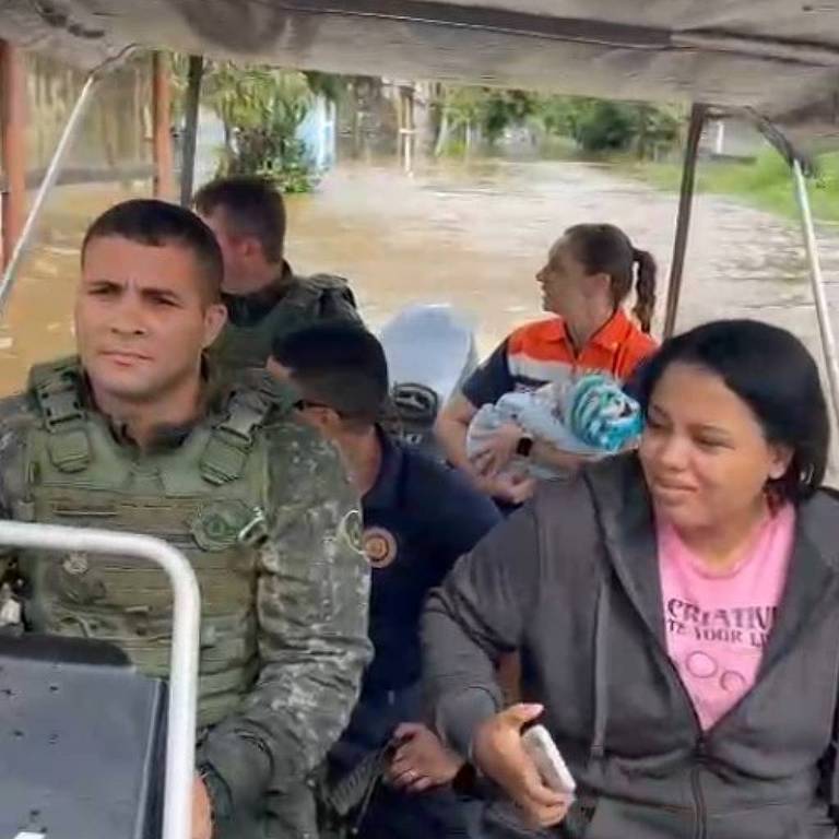 A imagem mostra um grupo de pessoas em um veículo de resgate. À frente， um homem vestido com uniforme militar está sentado ao volante. Ao lado dele， uma mulher com uma camiseta rosa parece estar segurando algo. Atrás deles， há várias crianças e um adulto， todos parecendo atentos à situação. O ambiente ao fundo sugere um cenário de emergência， possivelmente relacionado a inundações.