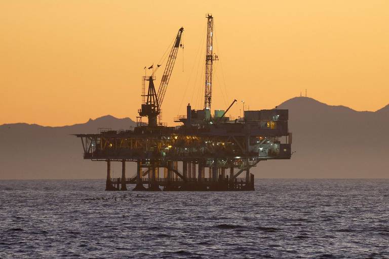 A imagem mostra uma plataforma de perfuração de petróleo localizada no mar， com guindastes visíveis. O céu está iluminado por um pôr do sol laranja， e ao fundo， há montanhas em silhueta.