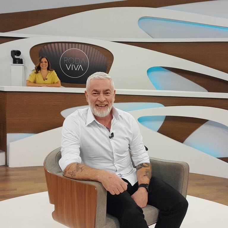 Um homem de cabelos grisalhos e barba, vestindo uma camisa branca, está sentado em uma poltrona em um estúdio de televisão. Ele sorri para a câmera. Ao fundo, uma mulher com cabelo longo e vestido amarelo está sentada em uma mesa de apresentação, com o logotipo 'RODA VIVA' visível. O cenário é moderno, com elementos em madeira e detalhes em azul.