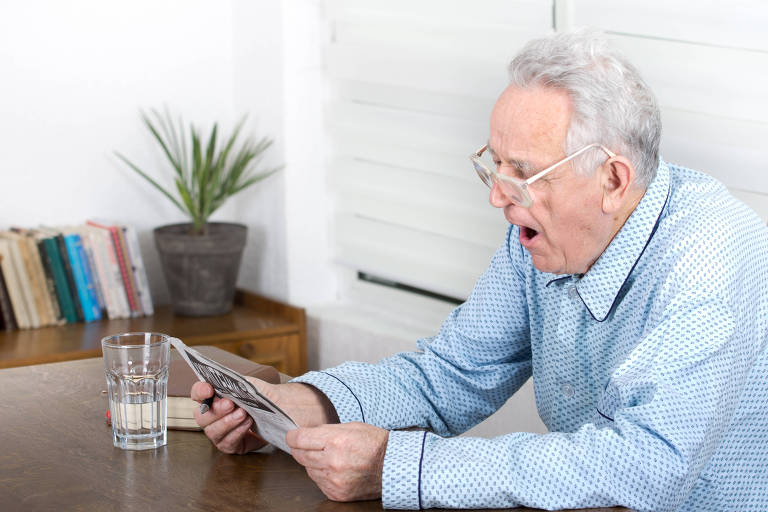 Um homem idoso está sentado à mesa， lendo um jornal com uma expressão de surpresa ou cansaço. Ele usa óculos e uma camisa azul com estampas. Ao seu lado， há um copo de água e uma planta em um vaso. Ao fundo， prateleiras com livros estão visíveis， e a luz entra pela janela com persianas.