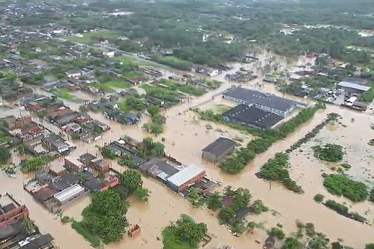 Imagem aérea de uma área urbana afetada por inundações， mostrando casas parcialmente submersas em água. A vegetação e as ruas estão cobertas， com algumas áreas de terra visíveis. O cenário é de grande extensão， com várias casas e um grande edifício em destaque， cercados por água.