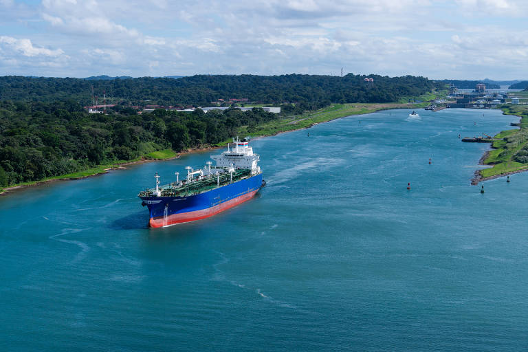 A imagem mostra um navio de carga navegando em um canal cercado por vegetação. O céu está parcialmente nublado e a água do canal é de um tom azul claro. À esquerda, há uma área de terra com árvores, enquanto à direita, o canal se estreita e se estende para o fundo da imagem.