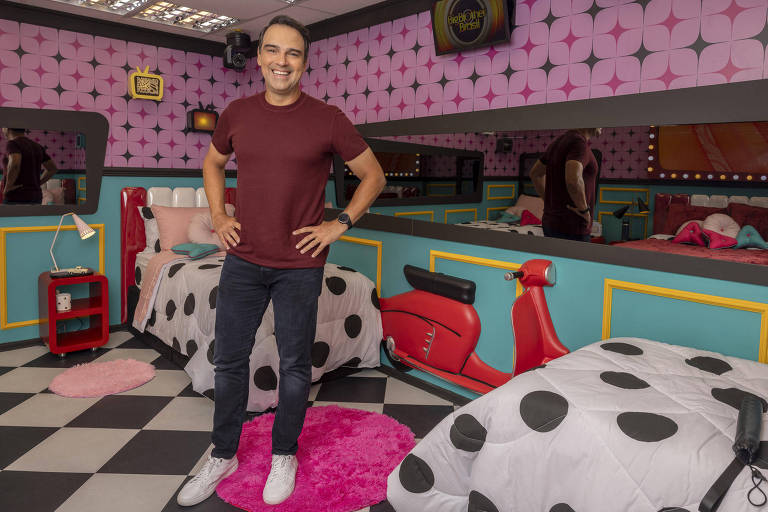 Um homem sorridente está posicionado em pé, com as mãos na cintura, em um ambiente colorido que parece ser o quarto do programa Big Brother Brasil. O fundo é decorado com paredes rosa e uma cama com colcha branca e bolinhas pretas. Há um espelho refletindo parte do ambiente e uma scooter vermelha decorativa ao lado de uma das camas. O chão é de ladrilhos em preto e branco, e há um tapete rosa em formato circular.