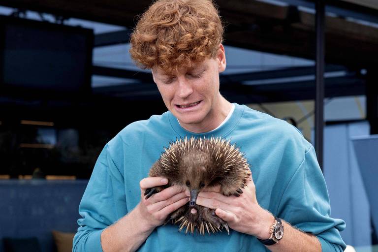 Um jovem com cabelo cacheado e ruivo está segurando uma equidna em suas mãos. Ele parece estar um pouco apreensivo ou emocionado, olhando para o animal. O fundo é desfocado, mas parece ser um ambiente interno com algumas estruturas visíveis.