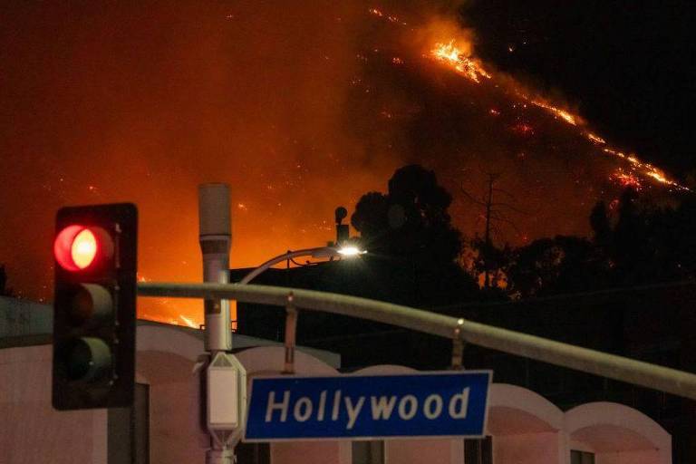 Placa de trânsito com localização de Hollywood em primeiro plano, com fogo ao fundo