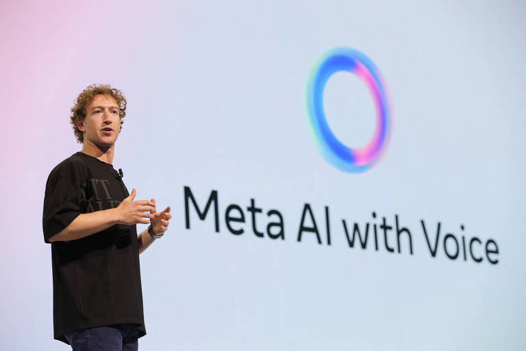 Imagem mostra Mark Zuckerberg， um homem que está em pé em um palco， gesticulando enquanto fala. Ao fundo， uma tela exibe o texto 039;Meta AI with Voice039; e um logotipo circular colorido. O apresentador está vestido com uma camiseta preta e parece estar em um evento de tecnologia.