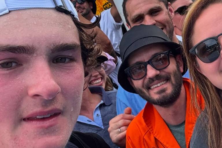 A imagem mostra um grupo de pessoas tirando uma selfie. No primeiro plano， um jovem com cabelo castanho e uma camiseta branca está sorrindo. Ao fundo， várias pessoas estão visíveis， incluindo um homem com um chapéu preto e óculos escuros， e outro usando uma camisa laranja. O ambiente parece ser um evento ao ar livre， com pessoas animadas ao redor.