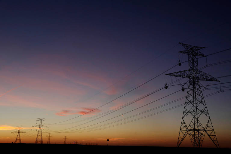 A imagem mostra várias torres de transmissão de energia elétrica contra um céu ao entardecer， com tons de azul， rosa e laranja. As torres estão localizadas em um campo aberto， e os fios de alta tensão se estendem entre elas.