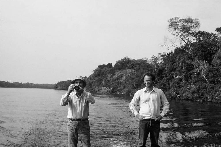 homem mais baixo fotografa em direção à câmera enquanto homem mais novo， à direita， põe as mãos na cintura sorrindo. ambos estão em frente a um rio com a floresta ao fundo. a foto está em preto e branco