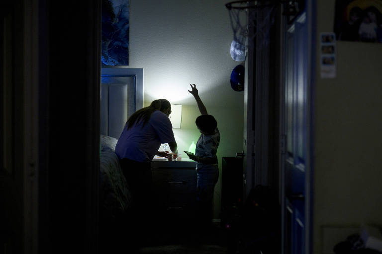 A imagem mostra duas silhuetas em um corredor escuro， onde uma pessoa adulta e uma criança estão próximas a uma mesa de cabeceira iluminada por uma lâmpada. A criança está levantando a mão， enquanto a pessoa adulta parece estar interagindo com ela. O ambiente é pouco iluminado， com paredes claras ao fundo.