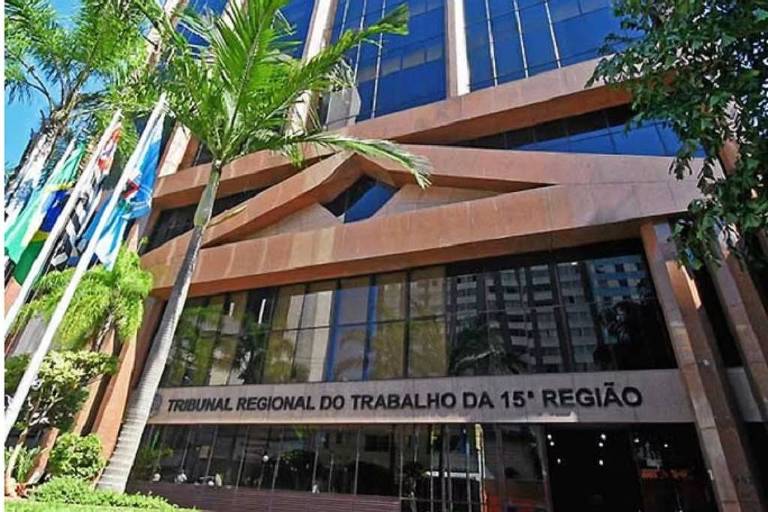 A imagem mostra a fachada do Tribunal Regional do Trabalho da 1ª Região. O edifício é moderno， com grandes janelas e uma entrada destacada. Há palmeiras ao lado e bandeiras visíveis na parte inferior da imagem. O nome do tribunal está escrito em letras grandes na parte inferior da fachada.