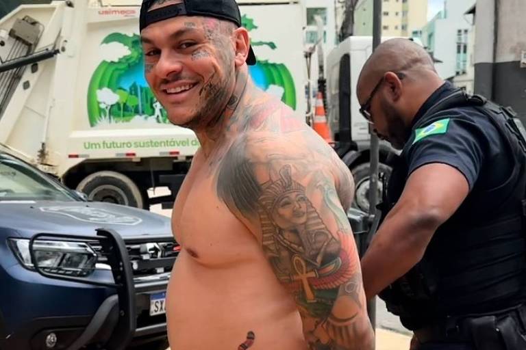 Um homem com tatuagens visíveis no corpo, incluindo uma grande tatuagem de uma figura egípcia no braço e uma cobra na barriga, está sorrindo enquanto é abordado por um policial. Ele está sem camisa e vestindo um short listrado azul e branco. Ao fundo, há um caminhão de lixo e edifícios. O policial está colocando algemas no homem.