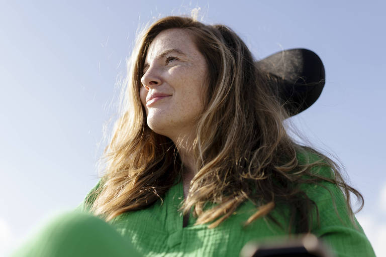 A imagem mostra uma mulher com cabelo longo e solto， usando uma roupa verde. Ela está sentada em uma cadeira de rodas， olhando para o horizonte com uma expressão serena. O céu está claro e azul ao fundo.