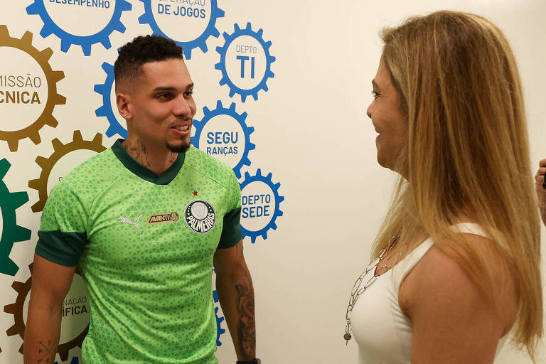 A imagem mostra um jogador de futebol usando uma camisa verde com detalhes em preto e o emblema do Palmeiras. Ele está sorrindo e conversando com uma mulher de cabelo longo e loiro， que usa uma blusa branca. Ao fundo， há uma parede decorada com engrenagens coloridas e textos como 039;COMISSÃO TÉCNICA039;， 039;DEPARTAMENTO TI039; e 039;JOGADORES039;.