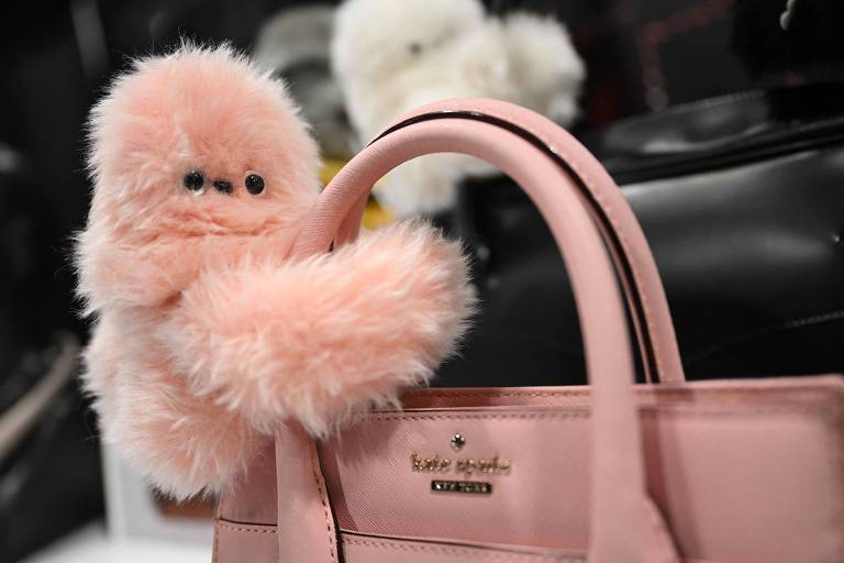 A imagem mostra um chaveiro de pelúcia rosa， com aparência de um pequeno monstro ou criatura， segurando a alça de uma bolsa rosa. A bolsa tem um design elegante e é da marca Kate Spade. Ao fundo， há outras bolsas e objetos desfocados.