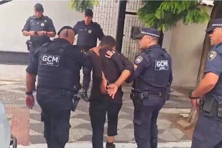 A imagem mostra uma operação policial em que dois policiais estão detendo um homem com as mãos algemadas. O homem está vestido de preto e é acompanhado por outros policiais que observam a situação. Ao fundo， há uma parede e algumas árvores. A cena parece ocorrer em uma área urbana.