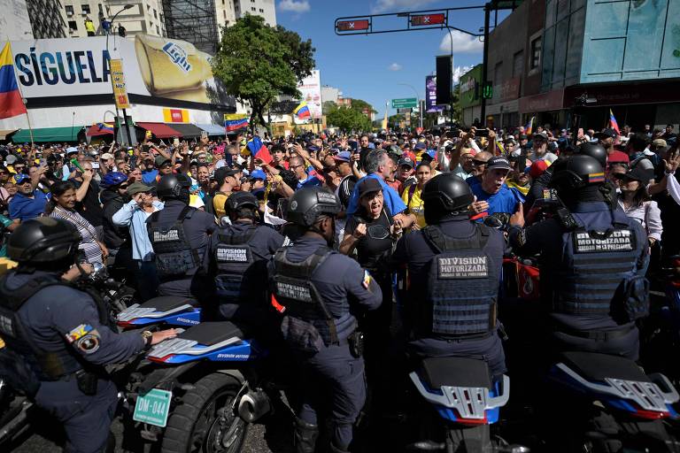 A imagem mostra uma grande multidão participando de um protesto em uma rua， com pessoas segurando bandeiras e cartazes. À frente da multidão， há policiais em motocicletas， vestidos com uniformes de segurança， formando uma barreira. Algumas pessoas na multidão parecem estar em um debate acalorado， enquanto outras estão levantando os braços. Ao fundo， há edifícios e uma grande faixa publicitária.
