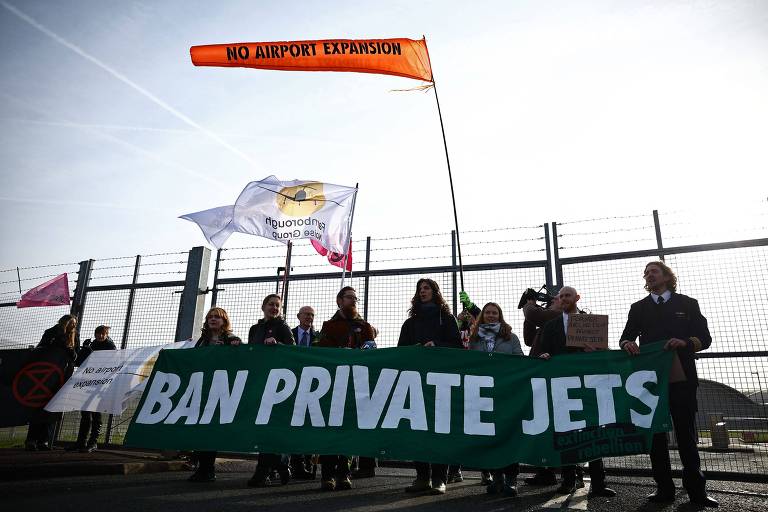 Um grupo de pessoas participa de um protesto segurando um grande banner verde com a frase 039;BAN PRIVATE JETS039;. Ao fundo， há uma cerca e uma bandeira laranja com a frase 039;NO MORE FLYING039;. O céu está nublado.