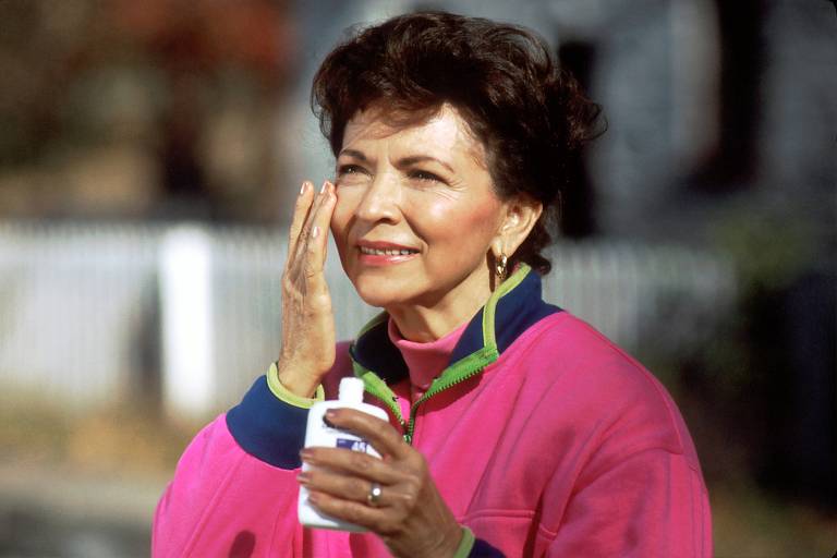 A imagem mostra uma mulher de pele clara， com cabelo castanho escuro e encaracolado， sorrindo enquanto aplica um creme na bochecha. Ela está vestindo uma jaqueta esportiva rosa com detalhes em azul e verde. A mulher segura um frasco de creme na mão. Ao fundo， há uma cerca branca e um ambiente ao ar livre.