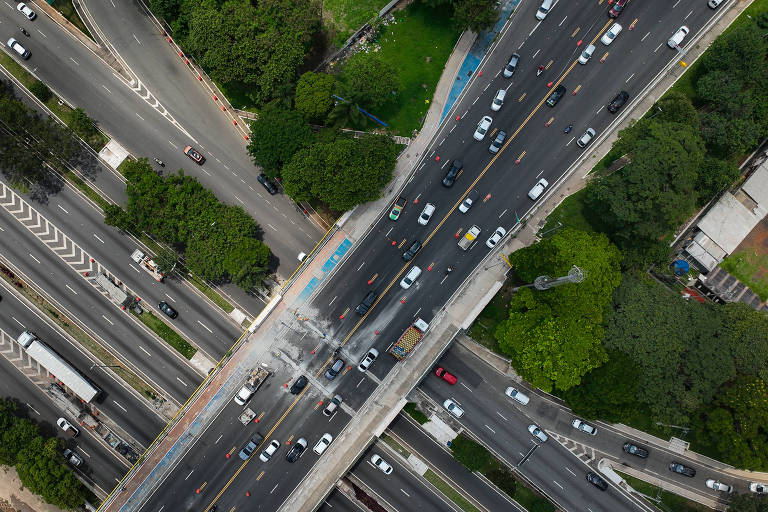 Imagem aérea de um complexo viário em uma área urbana， mostrando várias faixas de tráfego com carros em movimento. Há canteiros centrais com árvores e áreas verdes ao redor das estradas. Algumas partes da estrada estão em obras， com cones de sinalização visíveis. A imagem também revela a interseção de diferentes vias， incluindo uma ponte.