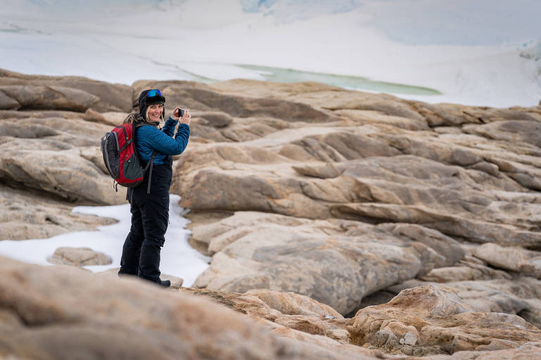 Uma mulher está em um terreno rochoso， sorrindo e segurando uma câmera. Ela usa uma jaqueta azul e uma mochila vermelha. O fundo apresenta uma paisagem de gelo e rochas， com algumas áreas cobertas de neve.